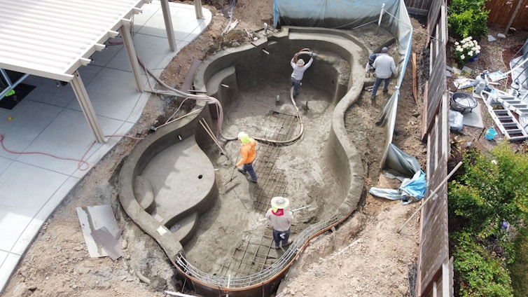 Three workers are actively involved in constructing a swimming pool. The pool is partially built and appears to be in the stages of concrete application. The surrounding area contains construction materials and a patio with a white pergola structure.