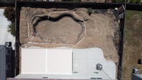 An aerial view of a backyard under construction, showcasing an outline of a swimming pool being built. The ground is excavated with concrete and steel reinforcements in place, indicating the preparation stages of a pool installation. A modern house with a light-colored roof and a concrete patio borders the left side of the construction area, and the backyard is enclosed by a wooden fence.