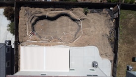 An aerial view of a backyard under construction, showcasing an outline of a swimming pool being built. The ground is excavated with concrete and steel reinforcements in place, indicating the preparation stages of a pool installation. A modern house with a light-colored roof and a concrete patio borders the left side of the construction area, and the backyard is enclosed by a wooden fence.