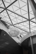 The interior of a modern architectural space is characterized by a large glass ceiling with triangular patterns. A prominent art installation resembling a large, intricate sculpture made of white materials hangs from the ceiling. The walls and structural elements are sleek with a matte black and white color scheme.