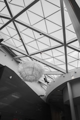 The interior of a modern architectural space is characterized by a large glass ceiling with triangular patterns. A prominent art installation resembling a large, intricate sculpture made of white materials hangs from the ceiling. The walls and structural elements are sleek with a matte black and white color scheme.