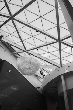 The interior of a modern architectural space is characterized by a large glass ceiling with triangular patterns. A prominent art installation resembling a large, intricate sculpture made of white materials hangs from the ceiling. The walls and structural elements are sleek with a matte black and white color scheme.