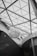 The interior of a modern architectural space is characterized by a large glass ceiling with triangular patterns. A prominent art installation resembling a large, intricate sculpture made of white materials hangs from the ceiling. The walls and structural elements are sleek with a matte black and white color scheme.