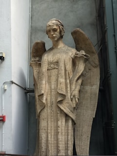 A tall stone statue of an angel stands against a gray wall. The angel is depicted with large, curved wings and wears a long, flowing robe. The expression on the angel's face is solemn and serene.