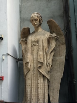 A tall stone statue of an angel stands against a gray wall. The angel is depicted with large, curved wings and wears a long, flowing robe. The expression on the angel's face is solemn and serene.