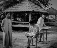 Three young men are in an outdoor setting with structures resembling small gazebos. One stands in traditional attire, while another sits casually on a chair and the third stands at a desk. The background is lush with trees and leaves.