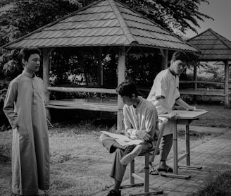 Three young men are in an outdoor setting with structures resembling small gazebos. One stands in traditional attire, while another sits casually on a chair and the third stands at a desk. The background is lush with trees and leaves.