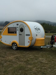 A small, modern teardrop trailer with a white body and bright yellow accents is parked on a grassy area. The trailer features a curved shape, a small window with a yellow frame, and a circular company logo displayed on its side. The trailer is attached to a vehicle that is partially visible.
