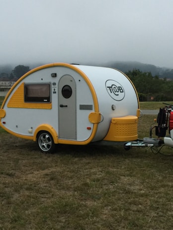 A small, modern teardrop trailer with a white body and bright yellow accents is parked on a grassy area. The trailer features a curved shape, a small window with a yellow frame, and a circular company logo displayed on its side. The trailer is attached to a vehicle that is partially visible.