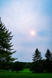 A serene outdoor landscape featuring lush green grass and several trees beneath a partly cloudy sky. The sun is visible through the clouds, casting a gentle light over the scene. In the distance, a group of people can be seen, possibly engaging in a leisure activity such as golfing.
