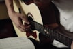 A person is playing an acoustic guitar, focusing on their hand positions. The musician is wearing a light-colored shirt. To the side, there is an open notebook with music notes and chord diagrams written on it.