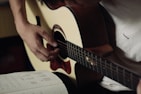 A person is playing an acoustic guitar, focusing on their hand positions. The musician is wearing a light-colored shirt. To the side, there is an open notebook with music notes and chord diagrams written on it.