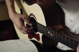 A person is playing an acoustic guitar, focusing on their hand positions. The musician is wearing a light-colored shirt. To the side, there is an open notebook with music notes and chord diagrams written on it.