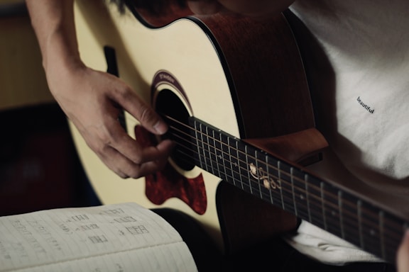 A person is playing an acoustic guitar, focusing on their hand positions. The musician is wearing a light-colored shirt. To the side, there is an open notebook with music notes and chord diagrams written on it.