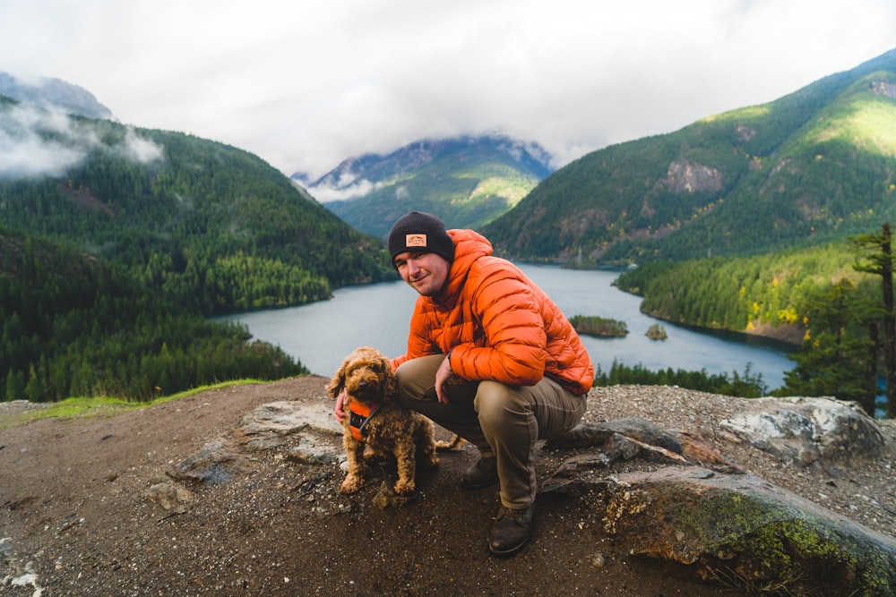 Mann in orangefarbenem Kapuzenpulli und schwarzer Hose sitzt tagsüber neben einem braunen Hund auf dem Felsen