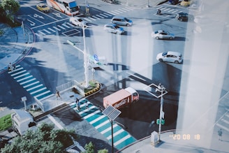 An urban intersection with several lanes of traffic, including a red delivery truck, cars, and a bus. Pedestrians are crossing the street on distinct zebra crossings, while some greenery and street signs are visible. The scene includes shadows cast by tall structures.