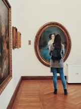A person stands in an art gallery, observing a large, ornately framed portrait on the wall. The portrait depicts a woman in a blue dress set against a dark background. The gallery has wooden flooring and additional paintings are displayed along the walls.