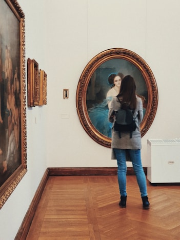 A person stands in an art gallery, observing a large, ornately framed portrait on the wall. The portrait depicts a woman in a blue dress set against a dark background. The gallery has wooden flooring and additional paintings are displayed along the walls.