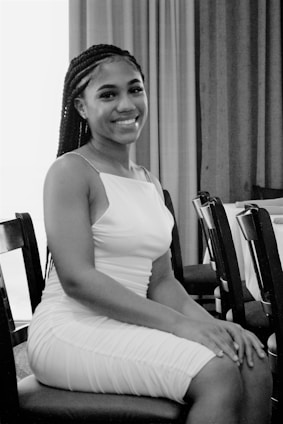A woman with braided hair is sitting on a chair in a setting that appears to be indoors. She is wearing a sleeveless white dress and smiling at the camera. The background consists of drawn curtains and additional chairs, suggesting a formal or semi-formal setting.