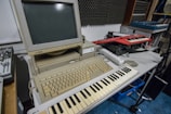 A vintage Atari computer setup is placed on a table, featuring a beige monitor and keyboard. In front of the computer is a MIDI keyboard. Additional electronic music equipment, such as another keyboard synthesizer, is seen on the side. The room has soundproofing panels on the wall, suggesting a music studio setting.