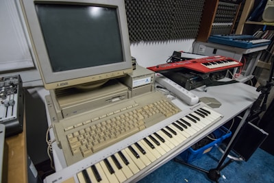 A vintage Atari computer setup is placed on a table, featuring a beige monitor and keyboard. In front of the computer is a MIDI keyboard. Additional electronic music equipment, such as another keyboard synthesizer, is seen on the side. The room has soundproofing panels on the wall, suggesting a music studio setting.