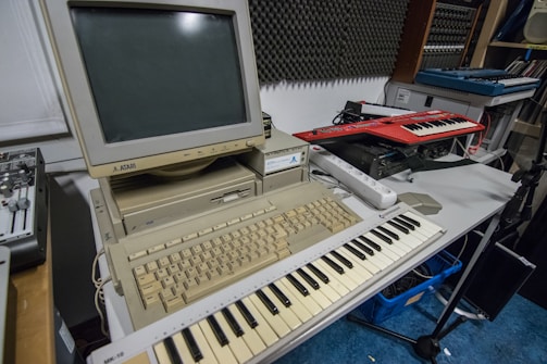 A vintage Atari computer setup is placed on a table, featuring a beige monitor and keyboard. In front of the computer is a MIDI keyboard. Additional electronic music equipment, such as another keyboard synthesizer, is seen on the side. The room has soundproofing panels on the wall, suggesting a music studio setting.