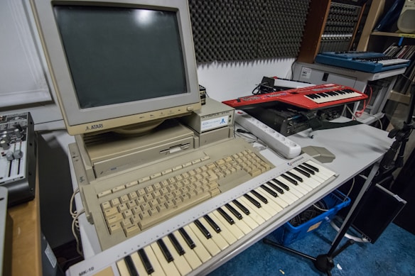 A vintage Atari computer setup is placed on a table, featuring a beige monitor and keyboard. In front of the computer is a MIDI keyboard. Additional electronic music equipment, such as another keyboard synthesizer, is seen on the side. The room has soundproofing panels on the wall, suggesting a music studio setting.