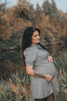 A pregnant woman stands in a natural setting with a calm expression and a hand resting on her belly. She's wearing a short-sleeved dress and has a tattoo on her forearm. The background features blurred autumn foliage.
