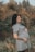 A pregnant woman stands in a natural setting with a calm expression and a hand resting on her belly. She's wearing a short-sleeved dress and has a tattoo on her forearm. The background features blurred autumn foliage.
