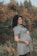 A pregnant woman stands in a natural setting with a calm expression and a hand resting on her belly. She's wearing a short-sleeved dress and has a tattoo on her forearm. The background features blurred autumn foliage.