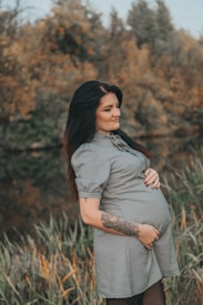 A pregnant woman stands in a natural setting with a calm expression and a hand resting on her belly. She's wearing a short-sleeved dress and has a tattoo on her forearm. The background features blurred autumn foliage.