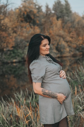 A pregnant woman stands in a natural setting with a calm expression and a hand resting on her belly. She's wearing a short-sleeved dress and has a tattoo on her forearm. The background features blurred autumn foliage.