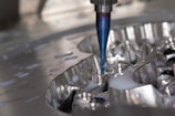 A close-up of a CNC milling machine cutting into a metal surface, with visible coolant fluid being applied around the area. The cutting tool is spinning, focusing on a detailed, reflective piece of metal.