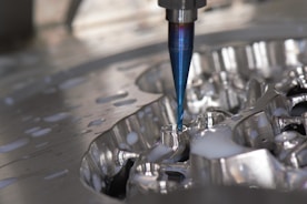 A close-up of a CNC milling machine cutting into a metal surface, with visible coolant fluid being applied around the area. The cutting tool is spinning, focusing on a detailed, reflective piece of metal.