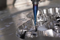 A close-up of a CNC milling machine cutting into a metal surface, with visible coolant fluid being applied around the area. The cutting tool is spinning, focusing on a detailed, reflective piece of metal.