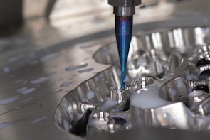 A close-up of a CNC milling machine cutting into a metal surface, with visible coolant fluid being applied around the area. The cutting tool is spinning, focusing on a detailed, reflective piece of metal.