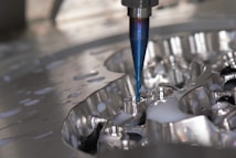 A close-up of a CNC milling machine cutting into a metal surface, with visible coolant fluid being applied around the area. The cutting tool is spinning, focusing on a detailed, reflective piece of metal.