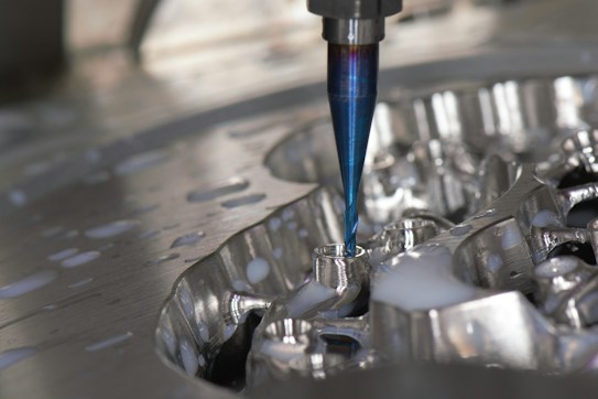 A close-up of a CNC milling machine cutting into a metal surface, with visible coolant fluid being applied around the area. The cutting tool is spinning, focusing on a detailed, reflective piece of metal.