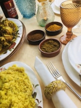 A table is elegantly set with various dishes and spices. There is a plate of chicken with yellow sauce, a bowl of yellow couscous, and small bowls containing different spices. Glasses of various colors and styles, along with a wine bottle, are also present. The tableware includes a napkin rolled in a woven holder, a fork, and a decorative plate.