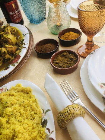 A table is elegantly set with various dishes and spices. There is a plate of chicken with yellow sauce, a bowl of yellow couscous, and small bowls containing different spices. Glasses of various colors and styles, along with a wine bottle, are also present. The tableware includes a napkin rolled in a woven holder, a fork, and a decorative plate.