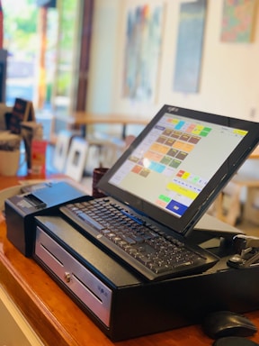 A point of sale system with a touchscreen monitor displaying a colorful software interface. The setup includes a keyboard, a cash drawer, a receipt printer, and a card reader. The background shows a blurred image of a bright, modern interior, likely a cafe or restaurant, with artwork on the walls and a large glass window providing a view of the outside.