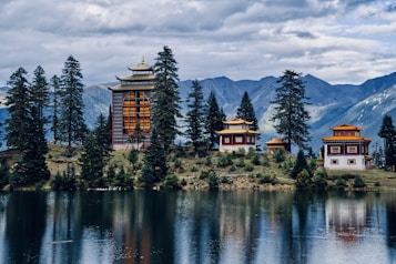 A serene lakeside scene featuring traditional Asian architecture amidst tall evergreen trees, with majestic mountains in the background under a cloudy sky. The buildings have ornate roofs with golden accents, enhancing the tranquil ambiance.