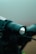 A close-up view of a handheld flashlight with droplets of water on its lens, positioned against a blurred background.
