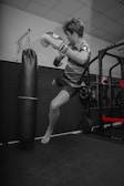 A person mid-air in a dynamic pose, wearing boxing gloves and kicking near a punching bag. The scene is set in a gym with a focus on physical activity, showing strength and motion.