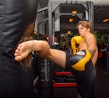 A person with yellow boxing gloves is performing a high kick against a black punching bag in a gym setting. The individual appears focused and is dressed in athletic shorts.