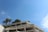 A modern white building with multiple balconies is adorned with lush green plants. Tall palm trees are visible against a clear blue sky, enhancing the serene and tropical atmosphere.