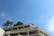 A modern white building with multiple balconies is adorned with lush green plants. Tall palm trees are visible against a clear blue sky, enhancing the serene and tropical atmosphere.