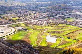 An aerial view of a sprawling suburban landscape featuring a large golf course with well-maintained greens, sand traps, and a central water reservoir. Surrounding the golf course are rows of residential houses and a network of roads. The background displays a dense area of buildings and greenery, showcasing a mixture of urban and natural elements.