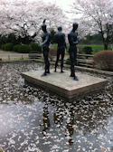 Three bronze statues stand on a stone platform surrounded by a reflective pool of water, scattered with cherry blossom petals. The background features lush greenery and cherry trees in full bloom, creating a serene and contemplative atmosphere.