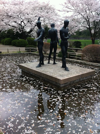Three bronze statues stand on a stone platform surrounded by a reflective pool of water, scattered with cherry blossom petals. The background features lush greenery and cherry trees in full bloom, creating a serene and contemplative atmosphere.